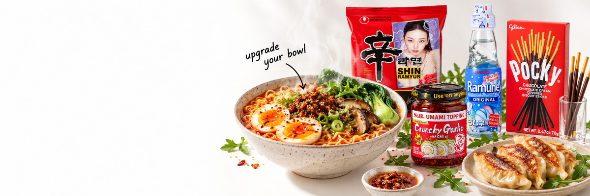 A steaming bowl of ramen surrounded by Shin Ramyun, chili crisp, Ramune soda, Pocky, and gyoza